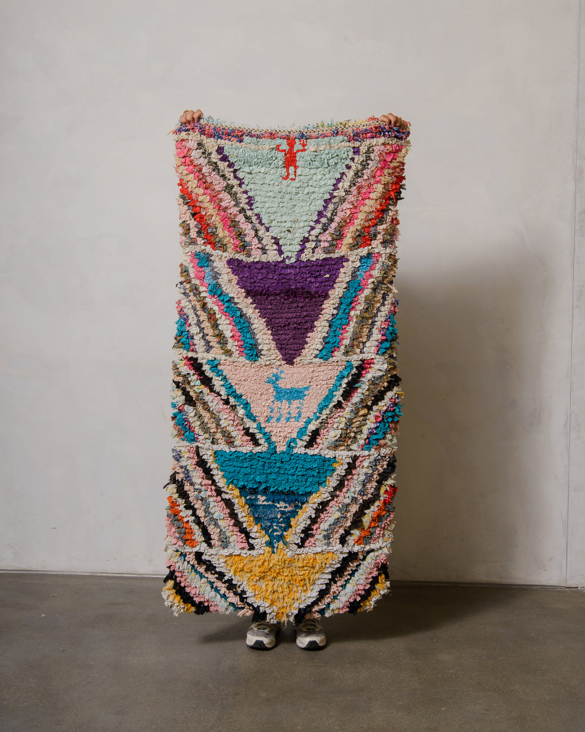 Berberlin boucherouite runner rug featuring blue, purple, yellow and pink, being held up against a white wall.